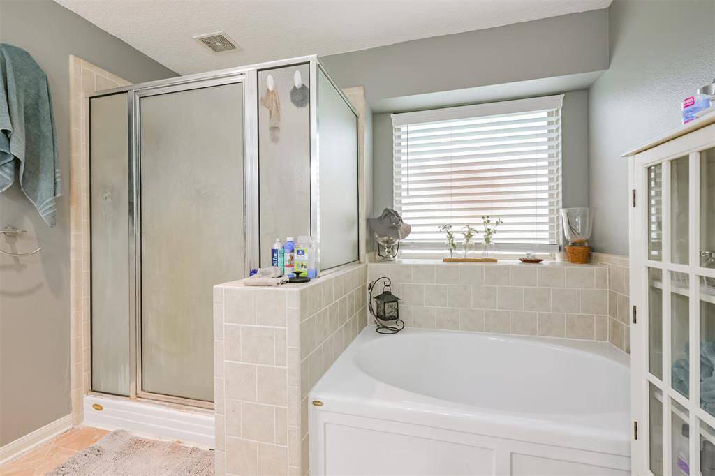 This is a well-lit bathroom featuring a large tub with tile surround, a separate frosted glass shower, and a window with blinds for natural light. There's a built-in storage cabinet to the right. The space has a clean, neutral color scheme.