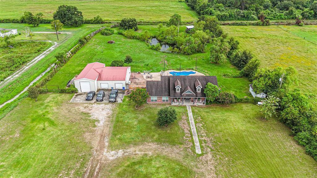 This is an aerial view of a spacious property featuring a large single-family home with a large workshop The home has a sizable backyard with an in-ground pool and is surrounded by expansive green fields, offering a sense of privacy and open space. There's also ample parking space available.