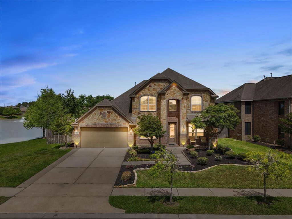 This photo showcases a beautiful two-story home with a stone and brick exterior. It features a well-maintained front yard with landscaped gardens and a spacious driveway leading to a two-car garage. The property is lake front w/ green belt on one side.