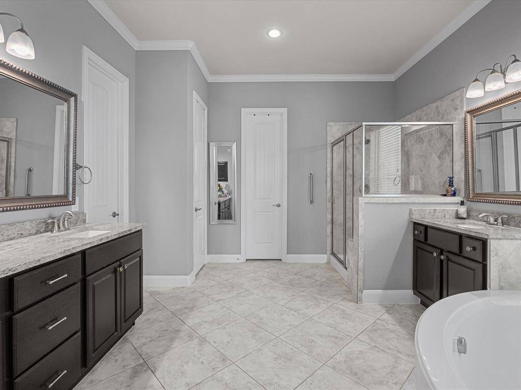 This spacious  primary bathroom features dual vanities with elegant granite countertops, a large glass-enclosed shower, and a separate soaking tub. The neutral tile flooring and soft gray walls create a calming atmosphere, complemented by modern lighting and ample storage.
