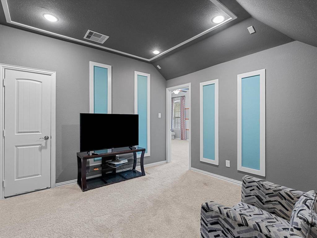 This cozy media room features a modern aesthetic with gray walls and blue accents, recessed lighting, and a plush carpet. It leads into the  adjoining game room through an open doorway.
