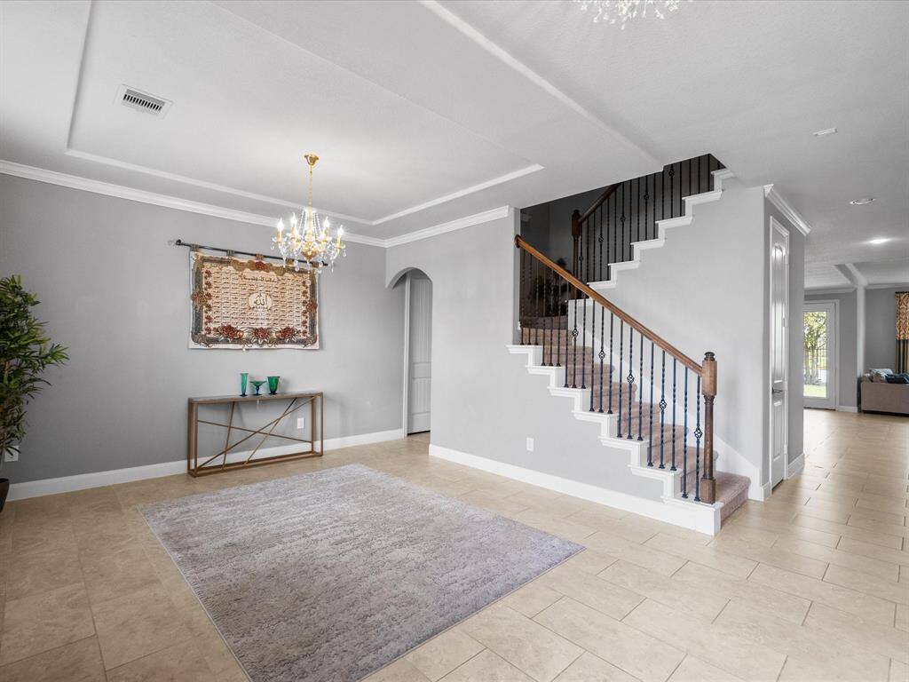 This elegant entryway features a stylish staircase with wrought iron railings, a decorative chandelier, and neutral tile flooring. An arched doorway and a light gray color palette enhance the spacious, inviting atmosphere.