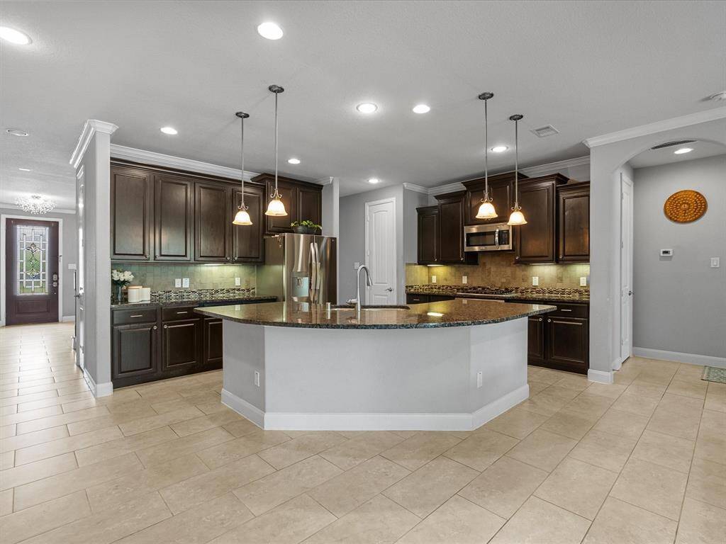 This spacious kitchen features dark wood cabinetry, a large island with pendant lighting, stainless steel appliances, and tile flooring. Adjacent is a cozy dining area, perfect for family meals.