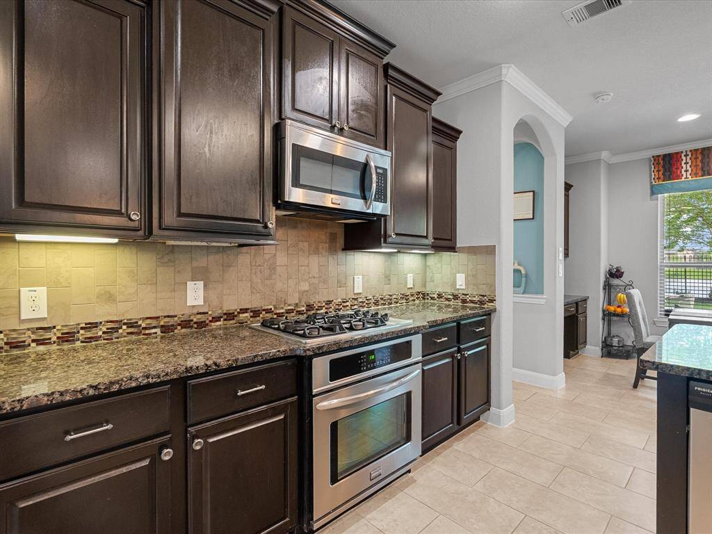 This spacious kitchen and dining area features modern appliances, a large island with pendant lighting, dark cabinetry, and a tile floor. It opens to a bright living space with large windows and a door leading to the outdoor area, perfect for entertaining and family gatherings.