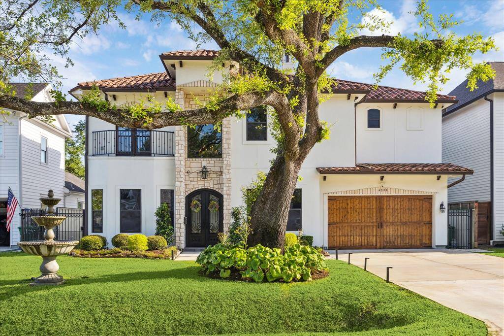 Elegant two-story home with a Mediterranean design, featuring a stucco exterior, clay tile roof, and arched windows. A large tree and a decorative fountain enhance the landscaped front yard. The property includes a spacious driveway leading to a wooden garage door.