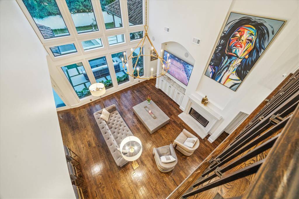 This is a stunning, open-concept living room with high ceilings and large windows that provide ample natural light. The space features elegant hardwood floors, modern furnishings, a fireplace. A stylish chandelier adds a touch of sophistication.