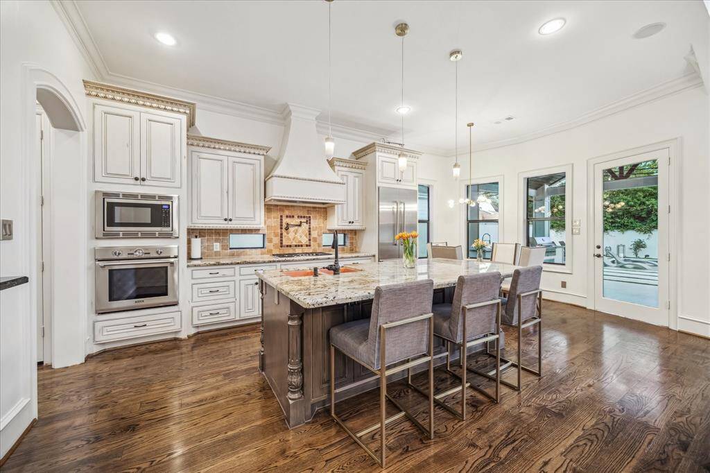 This kitchen features elegant white cabinetry, stainless steel appliances, and a spacious island with seating. It has hardwood floors and ample natural light from large windows and glass doors, creating a bright and inviting space.
