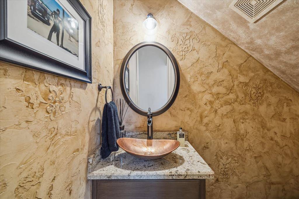 This cozy powder room features textured beige walls, a granite countertop, a stylish copper vessel sink, and a round mirror, creating a warm and inviting atmosphere.