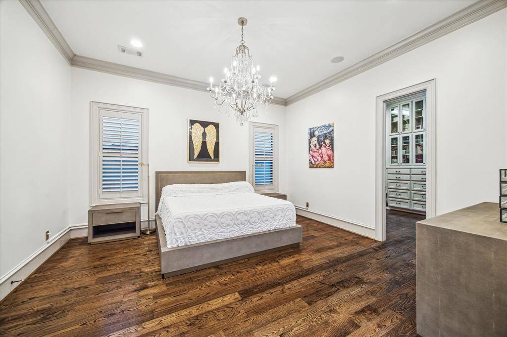 This master bedroom features elegant hardwood floors, a modern chandelier, and a neutral color palette. It includes a large bed, artwork on the walls, and an adjoining spacious walk-in closet with built-in cabinetry.