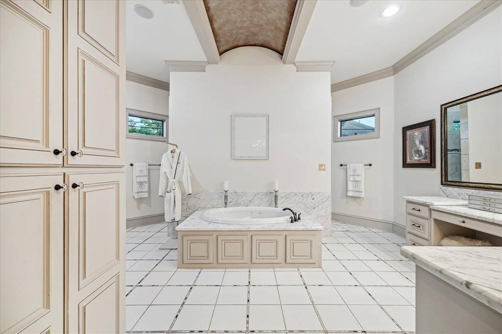 This elegant master bathroom features a luxurious central bathtub with a marble surround, ample cabinetry for storage, and a spacious vanity area. The neutral color palette, high ceiling, and natural light from small windows create an inviting and serene atmosphere.