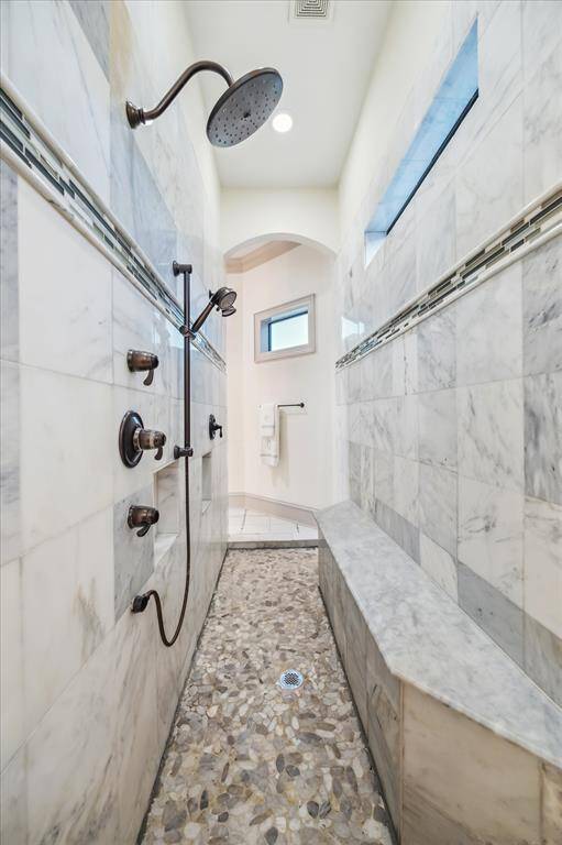 This photo showcases a luxurious, spa-like master shower with elegant marble tiles, a rainfall showerhead, and a pebble stone floor. The space features built-in seating and ample natural light, creating a serene and upscale bathroom experience.