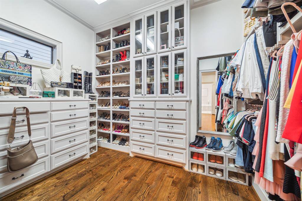 This photo showcases a corner of spacious walk-in closet featuring custom white cabinetry, ample shoe shelving, a variety of hanging spaces for clothing, and a full-length mirror. The hardwood floor adds warmth to the functional and stylish design.