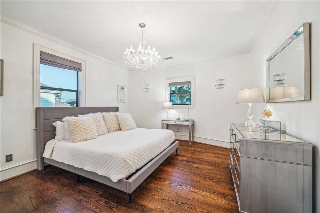 This bedroom features elegant hardwood flooring, a luxurious chandelier, and ample natural light from two windows. The room is tastefully furnished with a plush bed, mirrored dresser, and contemporary side tables, creating a sophisticated and serene atmosphere.