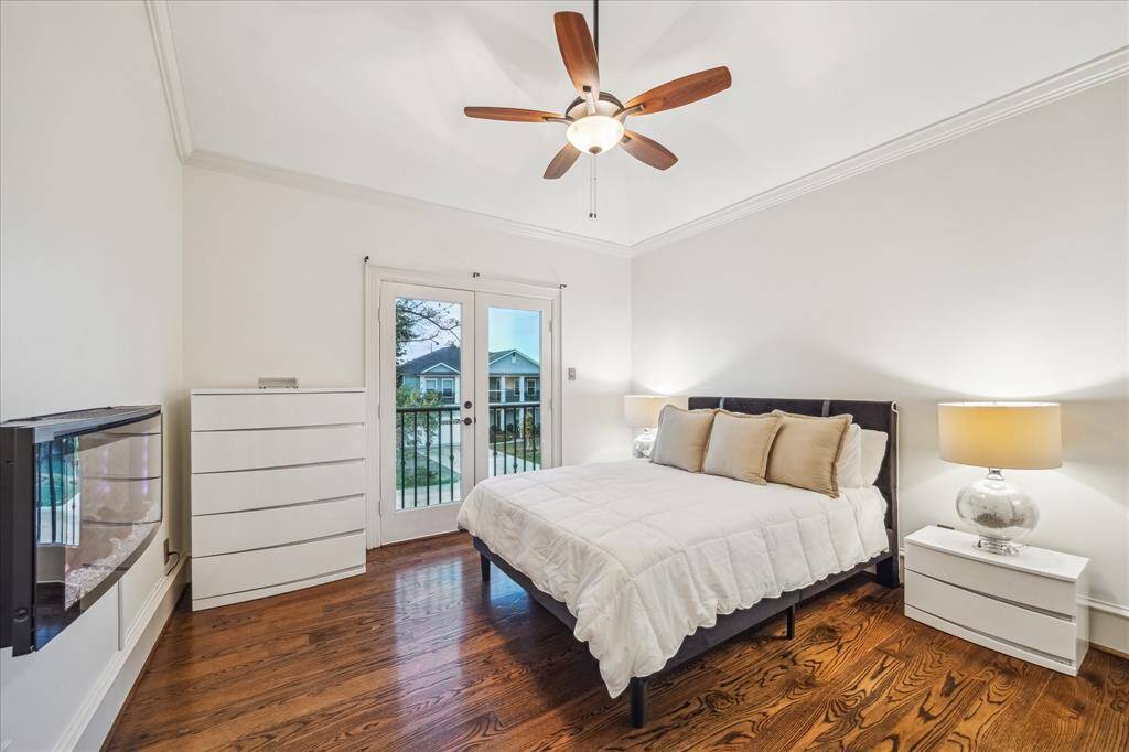 This bedroom features hardwood floors, a ceiling fan, and double doors opening to a balcony view. It includes modern furnishings with a cozy bed, nightstands, and a dresser, creating a bright and inviting space.