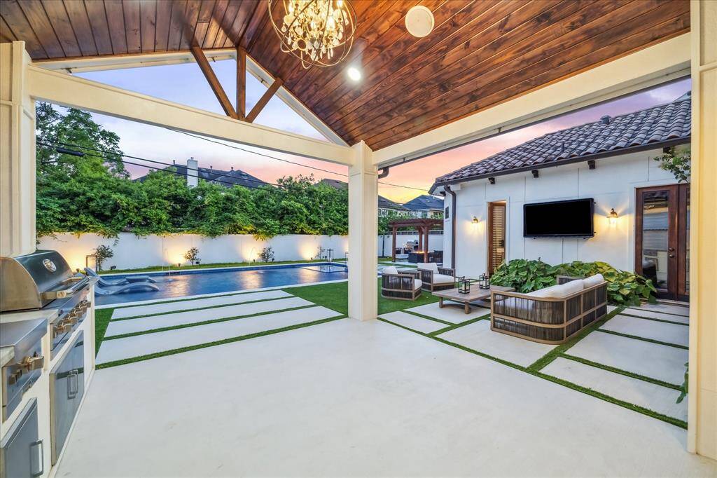 This outdoor space features a modern patio with a sleek BBQ area, a cozy seating arrangement, and a wall-mounted TV. It overlooks a spacious pool surrounded by lush greenery, creating an ideal setting for relaxation and entertaining.