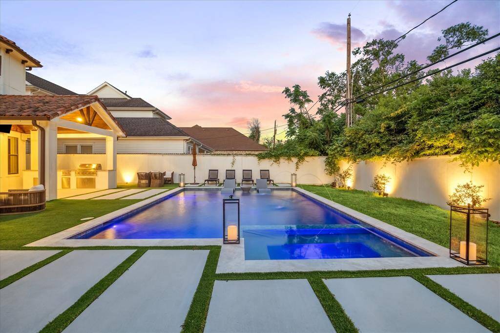 This photo showcases beautiful backyard with a modern swimming pool, surrounded by well-maintained greenery and ambient lighting. There's a covered outdoor kitchen and seating area, perfect for entertaining.