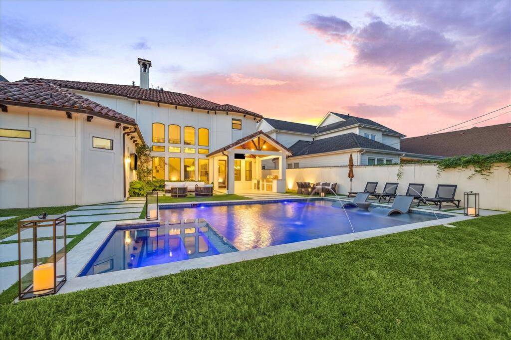 This stunning backyard features a modern pool with fountains, surrounded by lush grass and stylish lounge chairs. The home has a spacious patio area perfect for entertaining, and the backdrop of a beautiful sunset adds to the serene ambiance.