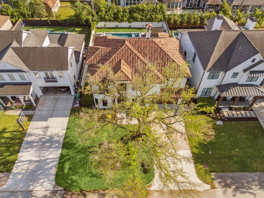 This aerial view showcases a charming home with a red-tiled clay roof nestled between two modern neighboring houses. The property features a spacious driveway and a well-maintained front yard. A large tree provides shade, and there's a swimming pool visible in the backyard, offering a great space for relaxation.