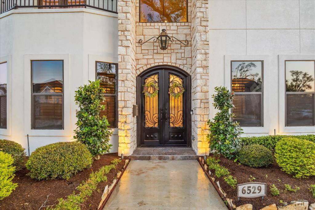 This photo showcases an elegant home entrance with a grand double door framed by stone accents. Beautiful landscaping with neatly trimmed bushes and plants enhances the curb appeal. Large windows flank the entrance, allowing natural light into the interior.