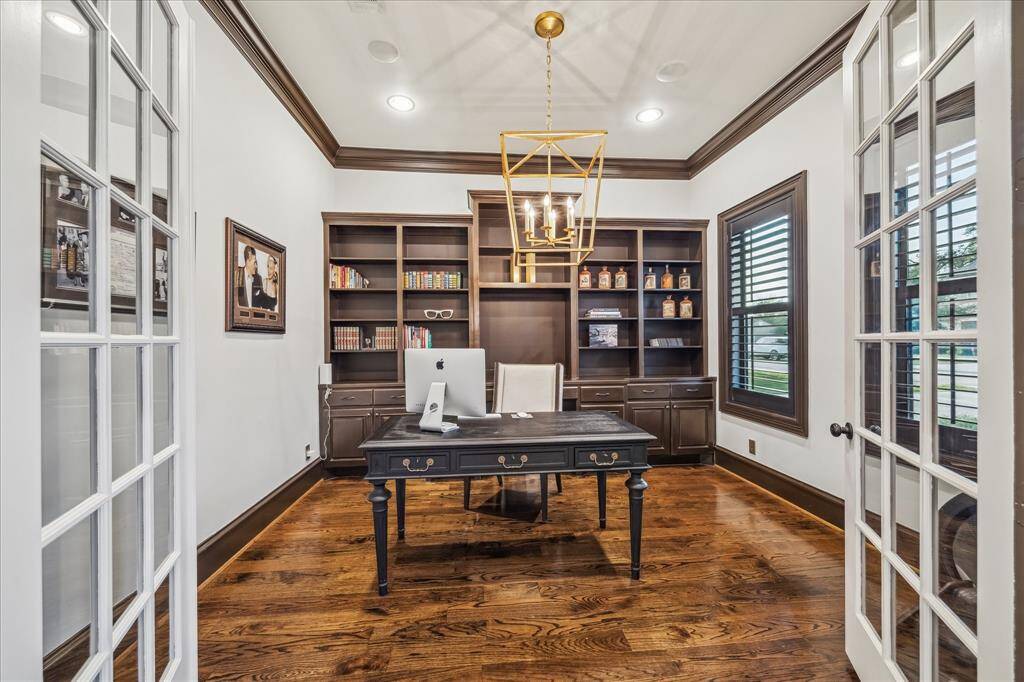 This elegant home office features rich wooden flooring, built-in bookshelves, and a classic desk. The room is brightened by large windows with plantation shutters and enhanced with a stylish chandelier. French doors add a sophisticated touch.