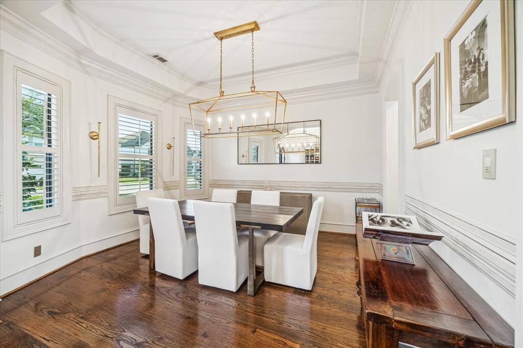 This dining room features elegant hardwood floors, a modern chandelier, and ample natural light from multiple windows with plantation shutters. The space is accented with tasteful wall art and a large mirror, creating a bright and inviting atmosphere perfect for entertaining.