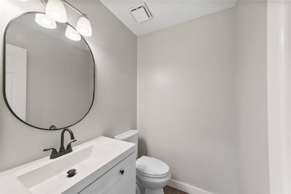 This half bathroom is tucked just off of the kitchen area and it boasts a low-profile white vanity with a cultured marble countertop and rectangular basin sink, modern black hardware, tile floors, and an oval-shaped framed mirror.