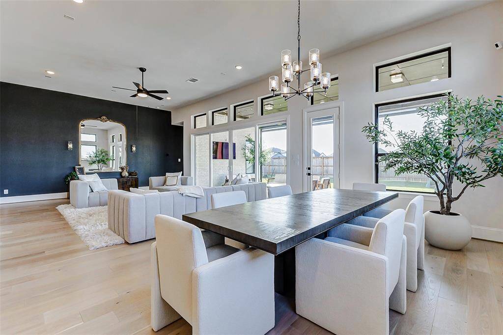 The dining area is highlighted by an elegant lighting fixture above the table, creating a focal point in the room. Expansive picture windows surround the space, flooding it with natural light and offering picturesque views. The open-concept layout further enhances the airy, inviting atmosphere.