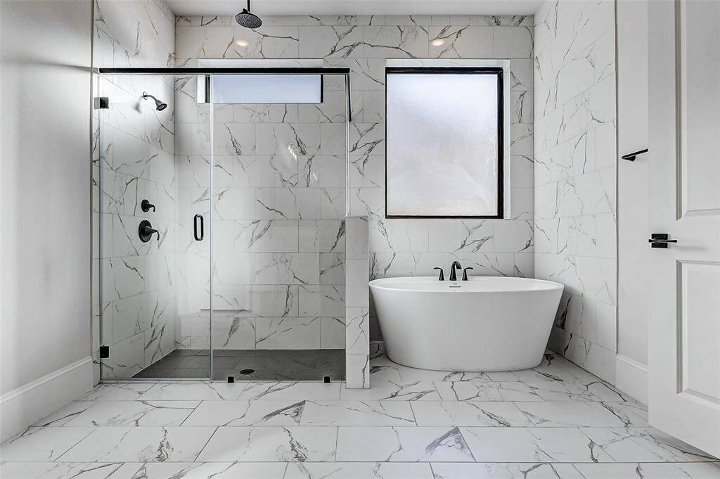 Step into this serene, spa-like retreat featuring floor-to-ceiling tile that adds a sleek, luxurious finish throughout. A freestanding tub sits gracefully beneath an obscure window, while the custom walk-in shower offers both a rain shower head and a standard shower head for a truly elevated experience.