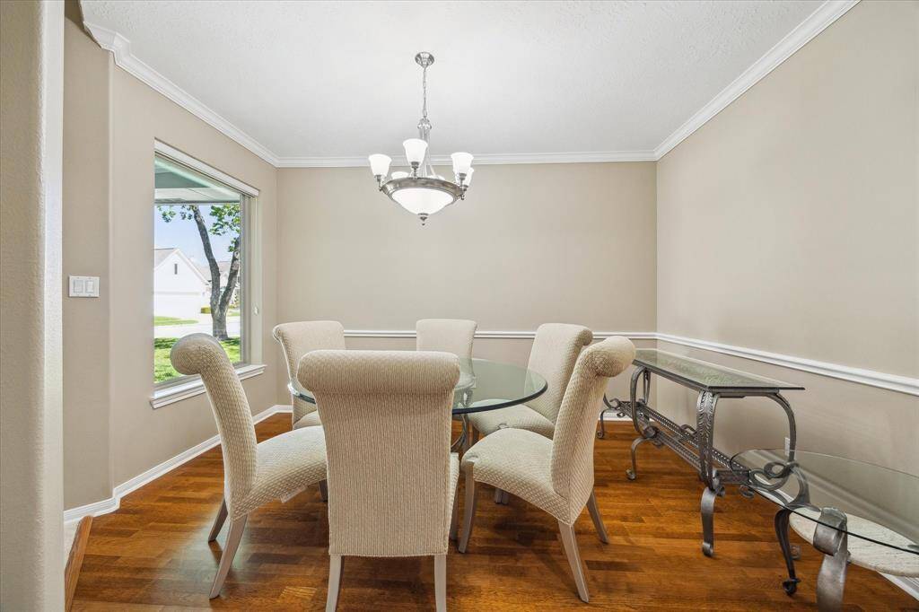 This dining room features warm wood-like flooring, neutral walls, and crown molding, with a large window offering natural light and a view of the outdoors. A stylish chandelier adds a touch of sophistication.