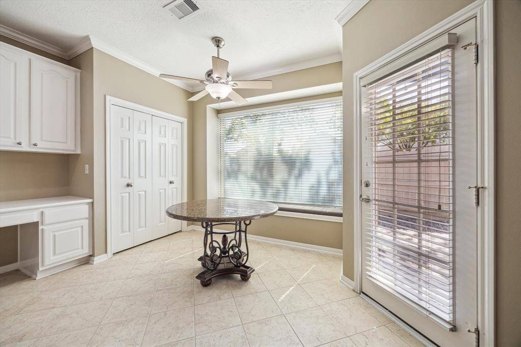 This bright breakfast area features a large window with blinds, a ceiling fan, and a space for working at home. It has tiled floors, built-in cabinetry, and a door leading to an outdoor space, offering a cozy and functional setting.