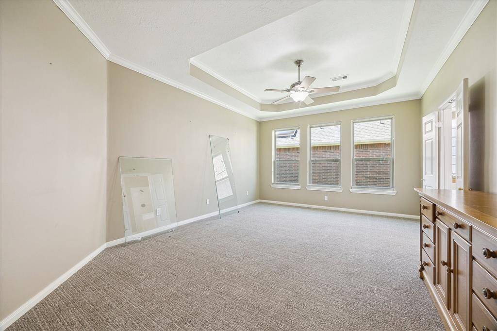 The spacious primary bedroom offers comfort and style with a tray ceiling, ceiling fan, and soft, neutral-toned carpeted flooring. Three large windows fill the room with natural light.