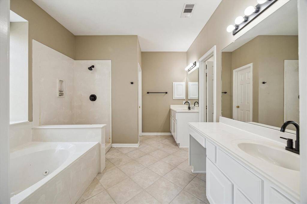 This spacious bathroom features dual vanities with sleek black fixtures, a large whirlpool tub, and a separate shower. It has a neutral color palette and ample natural light, creating a bright and inviting atmosphere. A frameless shower enclosure will be installed.