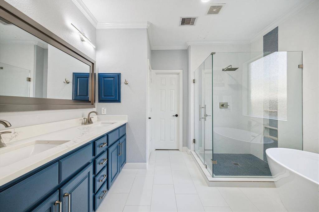 This spacious primary bathroom features a modern design with a large glass-enclosed shower, a sleek freestanding bathtub, and a double vanity with striking blue cabinets. It offers a clean, contemporary look with ample natural light.