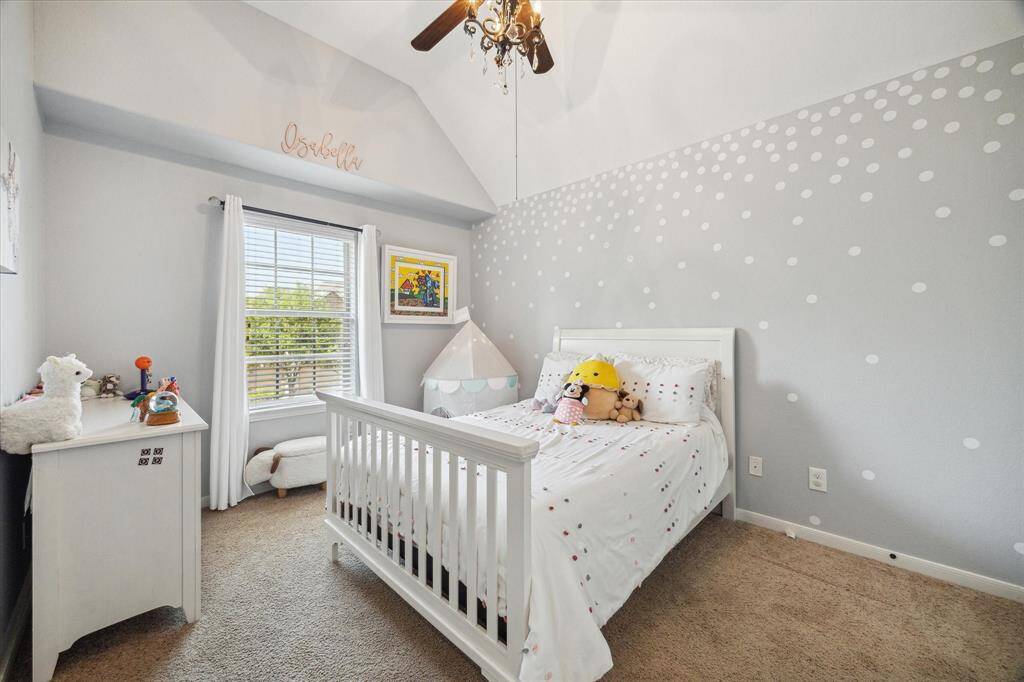 This is a cozy secondary bedroom with soft gray walls. It features plush carpet, and a ceiling fan with a light fixture. A window with white curtains provides natural light.