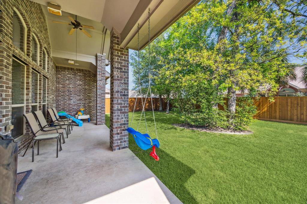 Charming backyard with a covered patio featuring seating and a ceiling fan. A swing and play area sit adjacent to a lush, fenced-in lawn, perfect for family activities and relaxation.