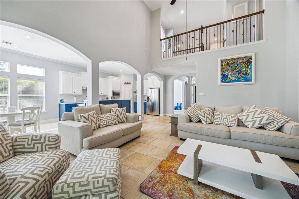 This spacious living area features high ceilings, abundant natural light, and an open floor plan. The room connects seamlessly to a stylish kitchen with white cabinets and blue accents. Perfect for comfortable living and entertaining.