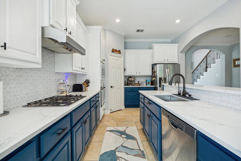 This kitchen features a modern design with a spacious layout, highlighted by white and blue cabinetry, a sleek tile backsplash, and stainless steel appliances. The room is bright with recessed lighting and includes a convenient double sink and cooktop, offering both functionality and style.
