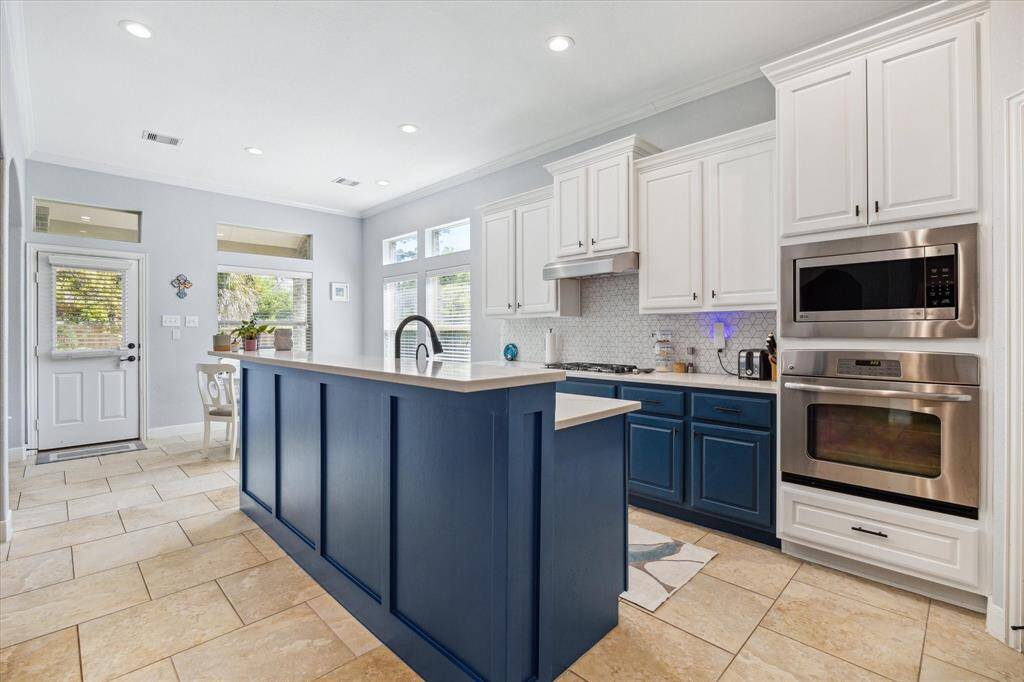 This kitchen features a modern design with white upper cabinets and blue lower cabinets. It includes stainless steel appliances, a central island with a sink, and tile flooring. Natural light fills the space through large windows, creating a bright and inviting atmosphere.
