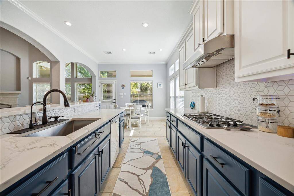 This kitchen features a modern design with white cabinetry, a sleek marble countertop, and a stylish hexagonal tile backsplash. It includes a built-in gas stove, deep sink, and ample natural light from large windows. The spacious layout flows into a breakfast area, creating an inviting space for cooking and entertaining.