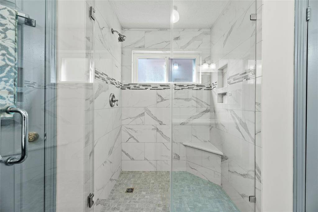 Primary bathroom walk-in shower featuring marble-style tile, a built-in corner seat, and frameless glass door.