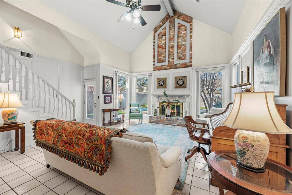 Alternate view of the living room featuring a high vaulted ceiling and a brick fireplace. Large windows provide ample natural light as well as allowing a great view of the patio and pool area.