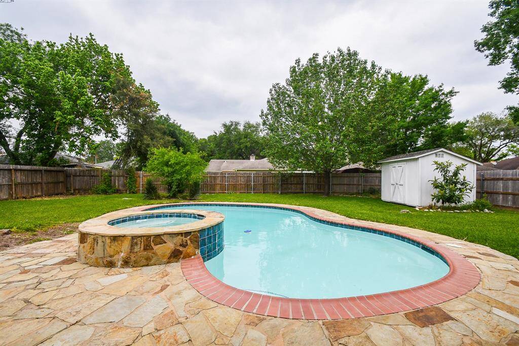 Spacious backyard featuring a luxurious pool with an attached spa, surrounded by a stone patio. Lush green lawn and mature trees provide privacy, complemented by a white storage shed. Perfect for outdoor relaxation and entertaining.