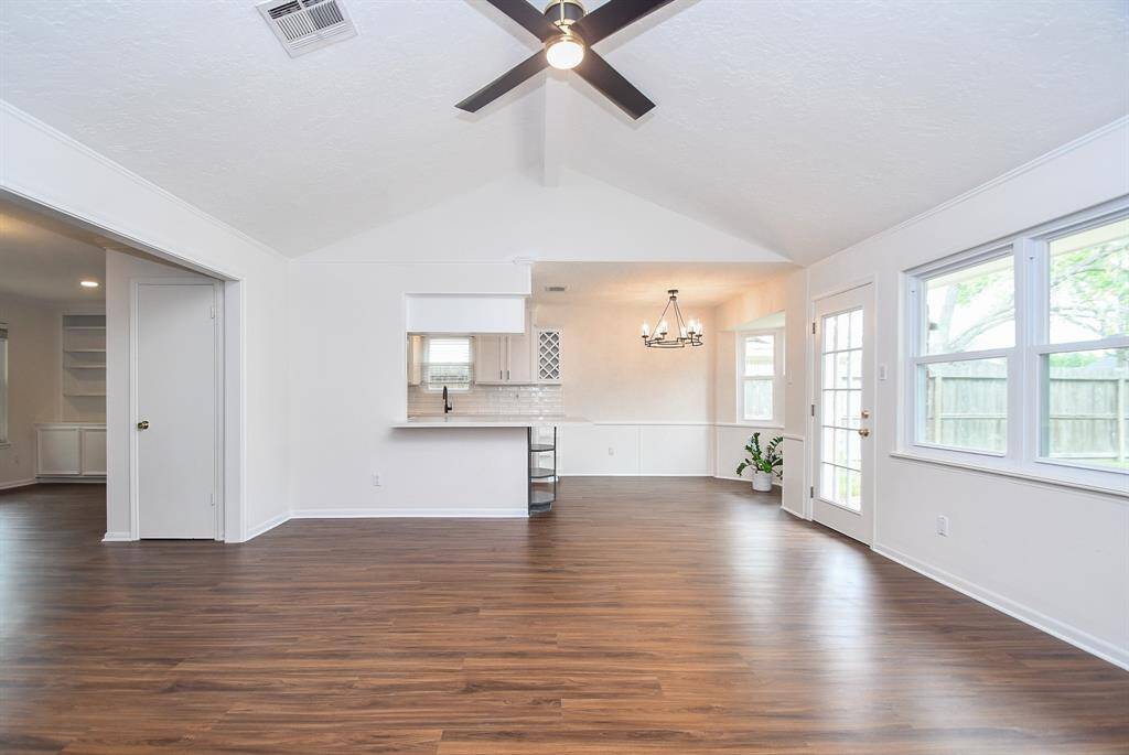 This photo showcases a spacious, open-concept living area with vaulted ceilings and abundant natural light. The room features warm floors, an energy efficient ceiling fan, and a modern chandelier. Large windows and a glass door lead to the backyard oasis, creating a bright and inviting atmosphere.