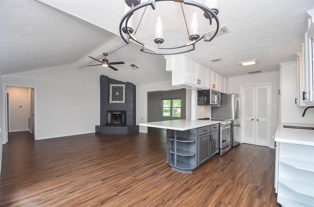 This spacious open-concept living area features a modern kitchen with sleek stainless steel appliances, ample cabinetry, and a large island. The room boasts beautiful wood flooring, a cozy brick fireplace, and a stylish ceiling fan, creating an inviting and functional space.
