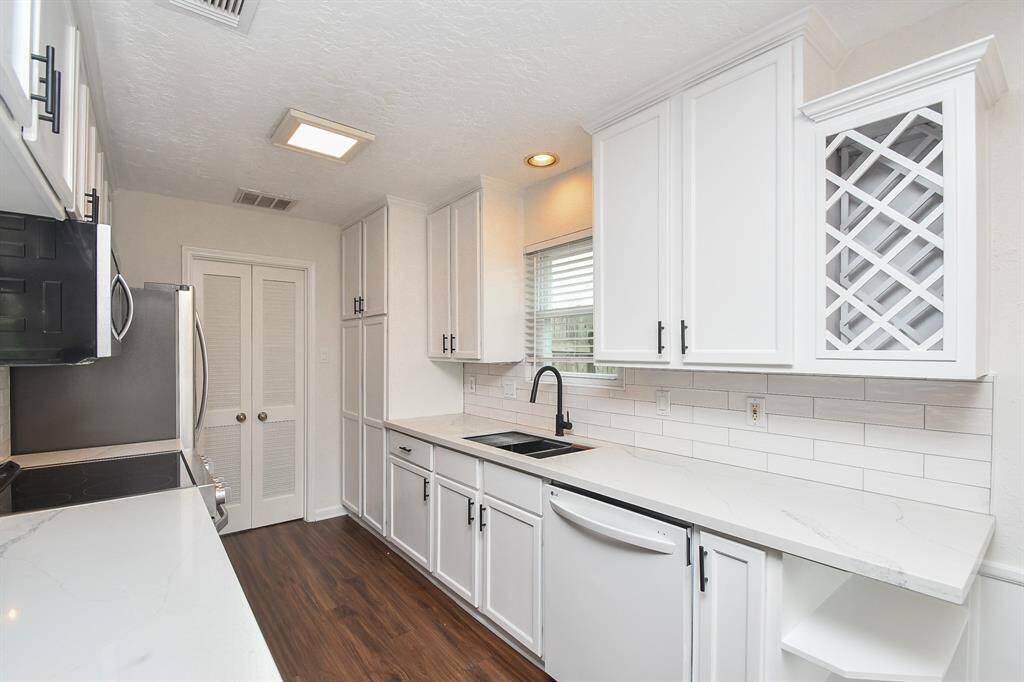 This kitchen features sleek white cabinetry, modern appliances, and a stylish black faucet. It offers ample counter space with a contemporary backsplash and a wine rack. The wood flooring adds warmth to the bright, streamlined design.