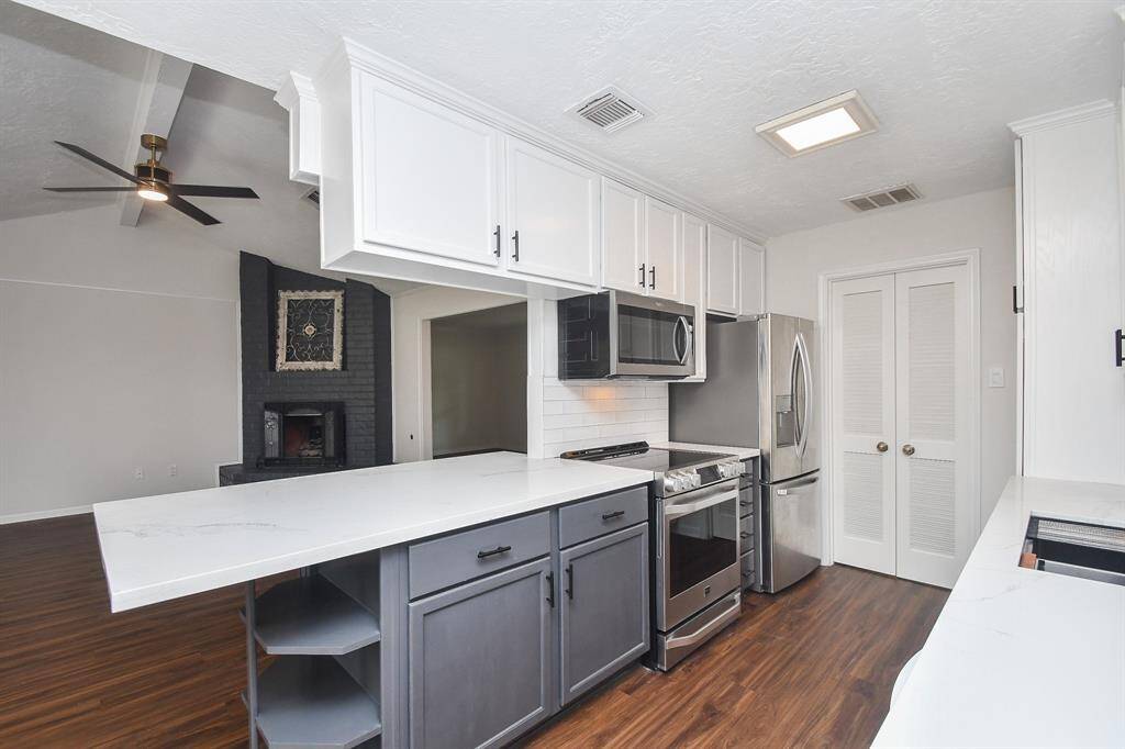 This modern kitchen features sleek stainless-steel appliances, white cabinetry, and a spacious island with a contrasting dark base. It opens into a living area with hardwood floors and a cozy, brick fireplace, creating a warm and inviting space.