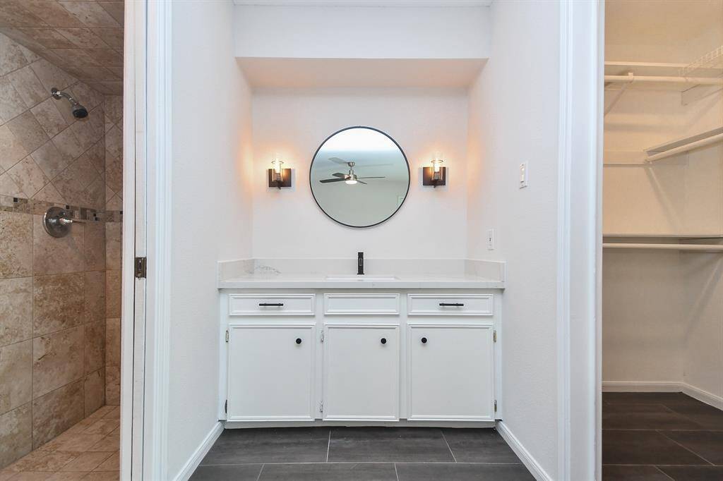 This bathroom features a sleek vanity with a round mirror and modern lighting. It has a tiled shower with a rainfall showerhead and an adjacent spacious walk-in closet which is one of two closets