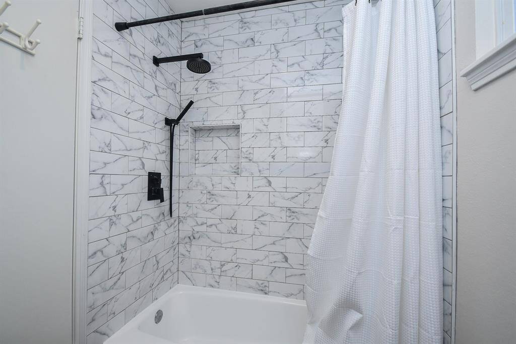 This bathroom features a sleek shower with modern black fixtures, a white tiled wall with marble pattern, a built-in niche for storage with ample space for toiletries in the cabinets behind.