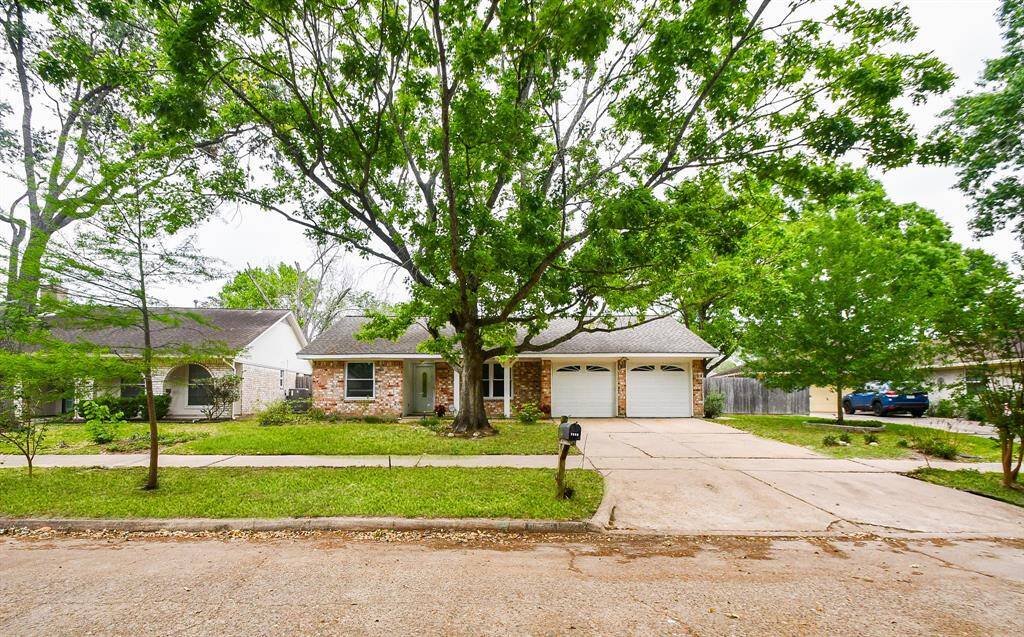 Charming single-story home with a brick facade, nestled under a large tree. Features a double garage and a spacious driveway, surrounded by lush greenery and a well-maintained lawn