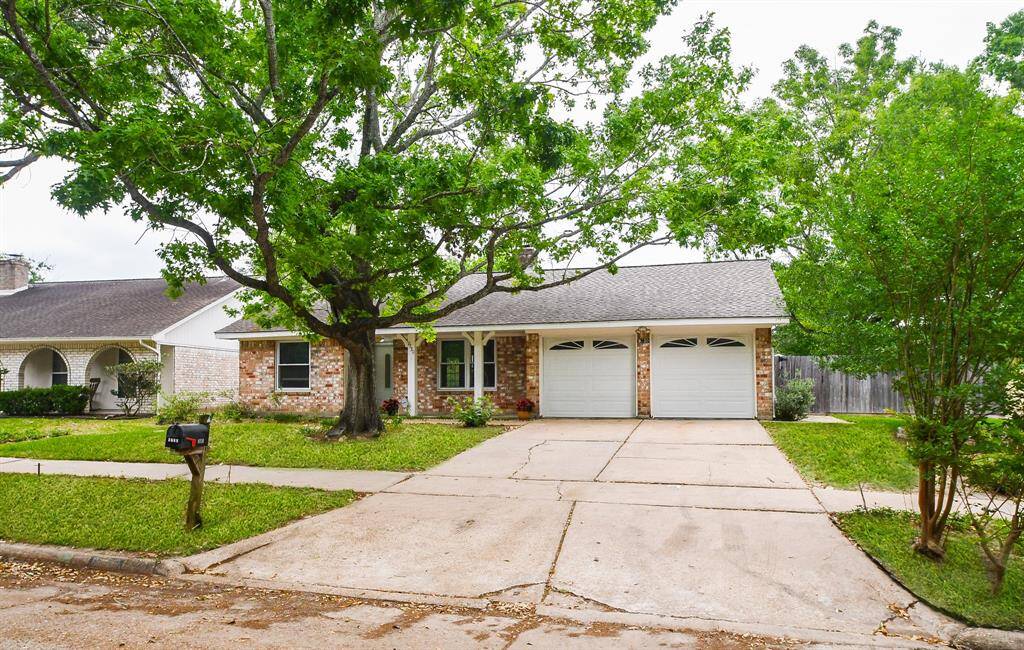 This charming single-story home features a brick exterior, a spacious two-car garage, and a welcoming front porch. A large tree provides shade over the driveway, and the front yard is neatly landscaped, offering great curb appeal.